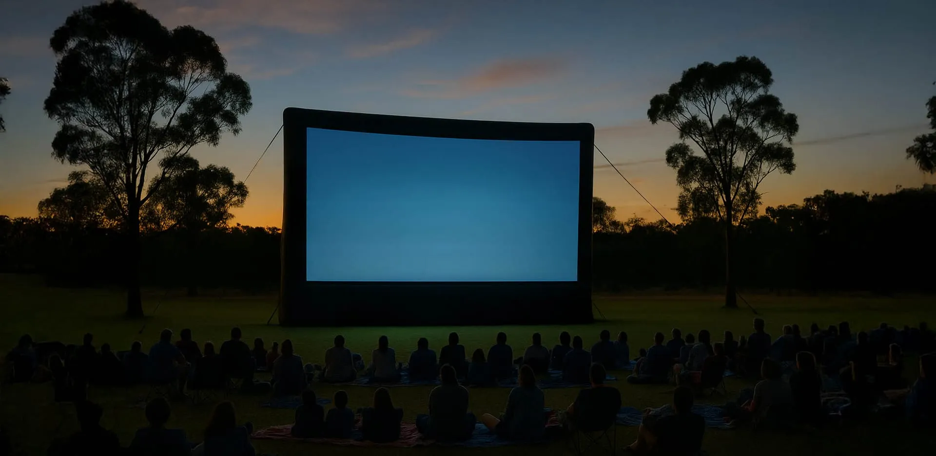 An outdoor cinema inflatable screen at dusk with a crowd seated on the grass