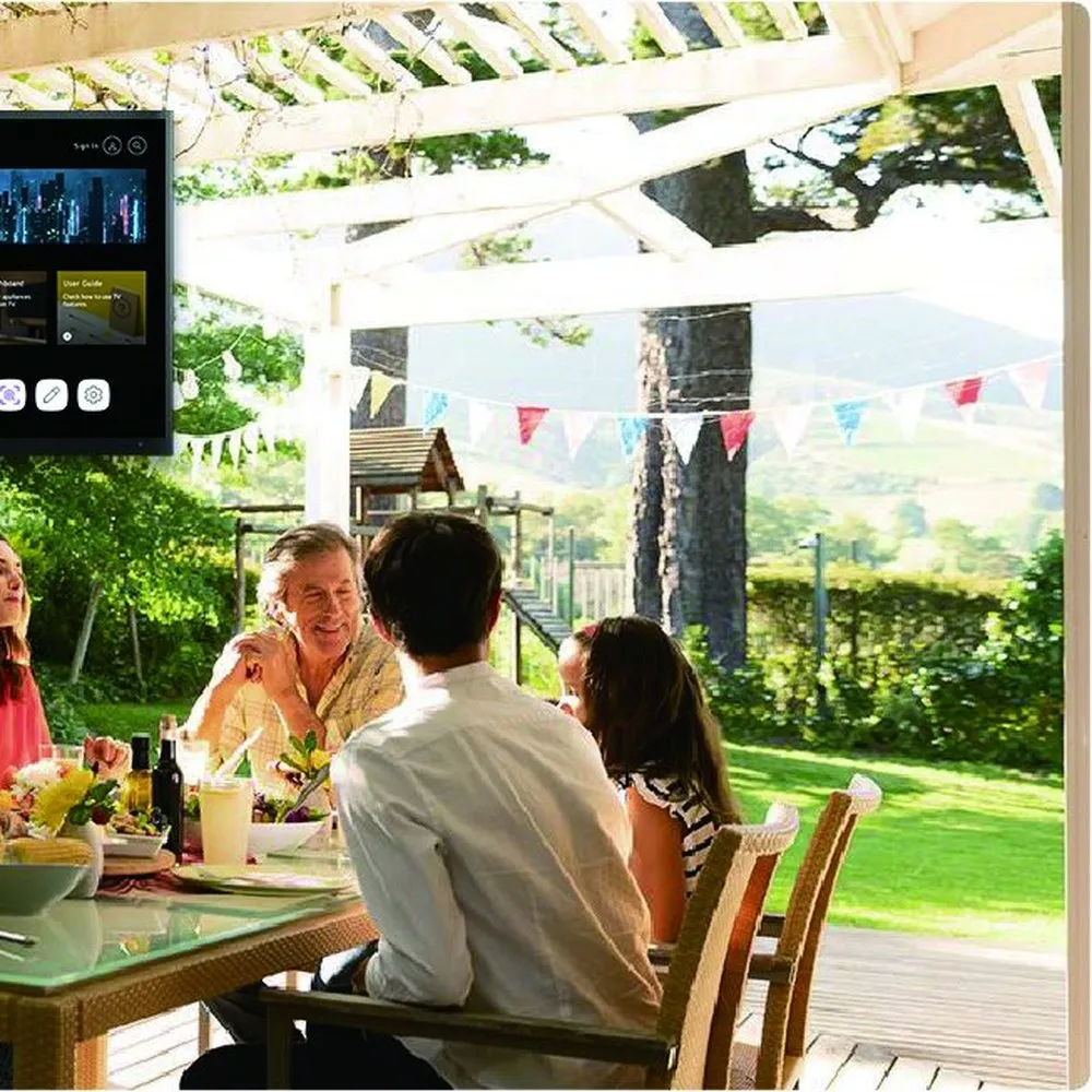 Diners seated at a long table in a covered outdoor dining area with a weatherproof television mounted on the wall