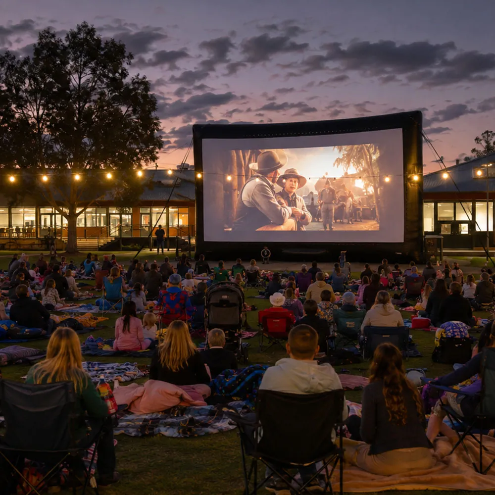A large public audience seated on grass watching an outdoor screen at night