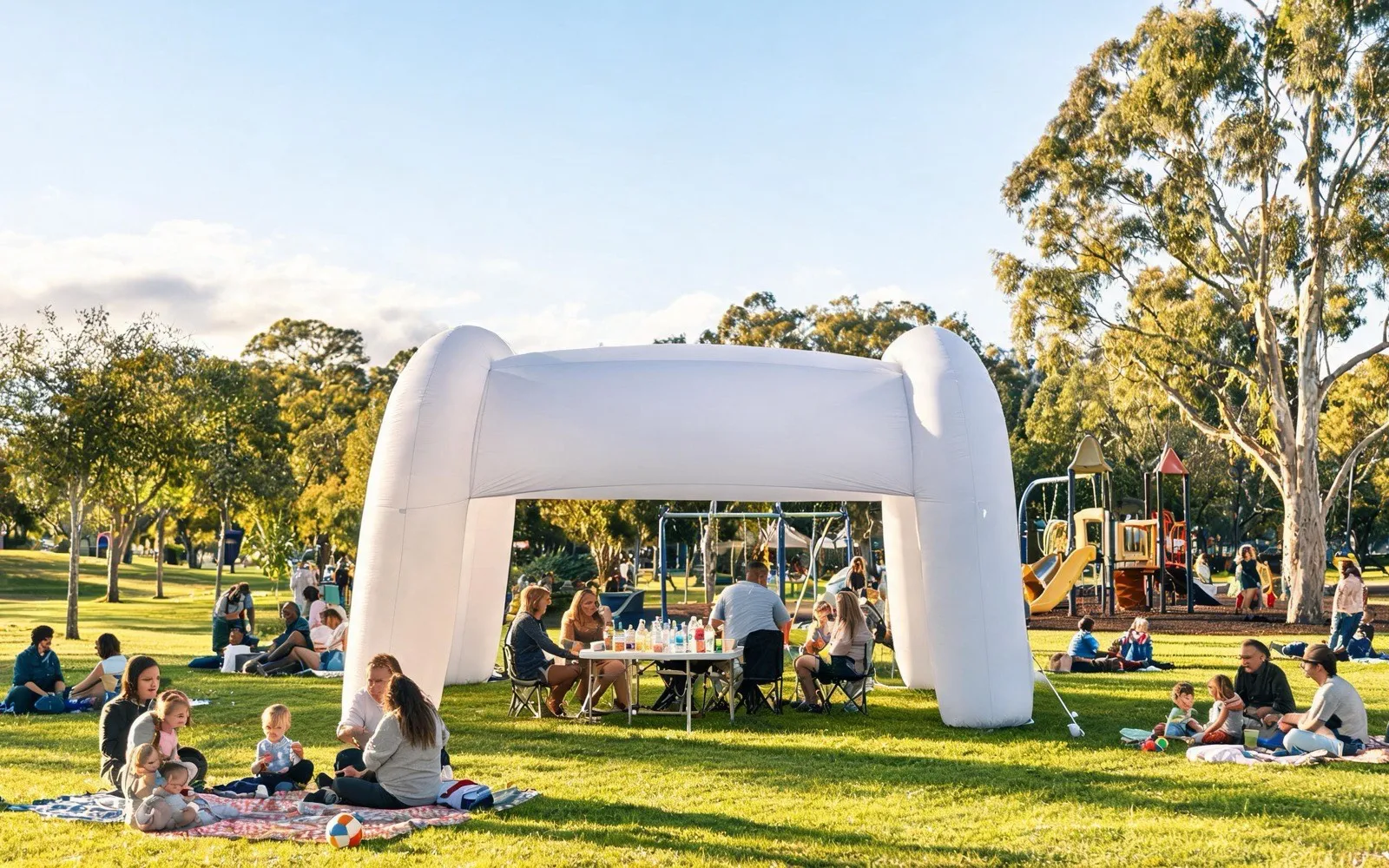 A white inflatable shelter in a suburban park at a community open day, with families on picnic rugs and a playground behind