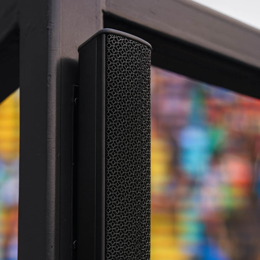 Black PA speaker with a textured surface against a colorful blurred background of an outdoor LED cinema display